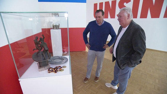 Jan Didjurgeit und Gerd Kische stehen vor einer Glasvitrine mit Trophäen. © Screenshot 