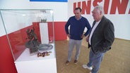 Jan Didjurgeit und Gerd Kische stehen vor einer Glasvitrine mit Trophäen. © Screenshot 