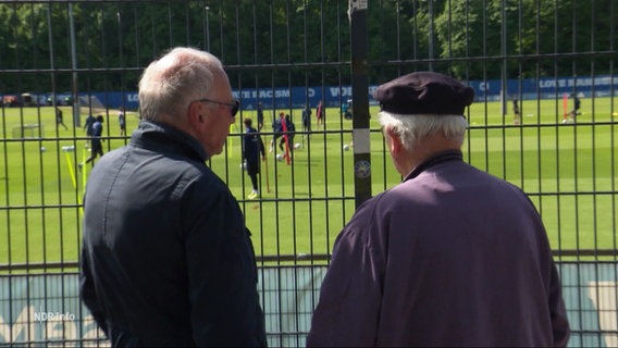 Zwei ältere Herren beobachten Fußballer hinter einem Zaun beim Training. © Screenshot 