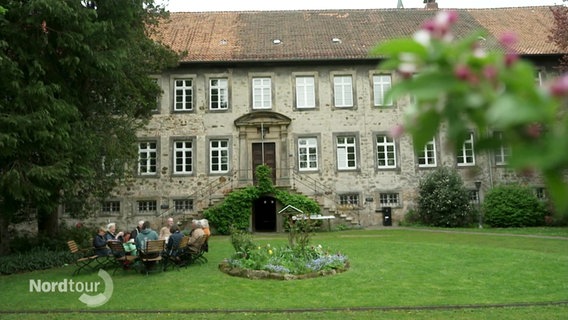 Eine Personengruppe sitzt an einem Tisch im Garten vor dem Kloster. © Screenshot 