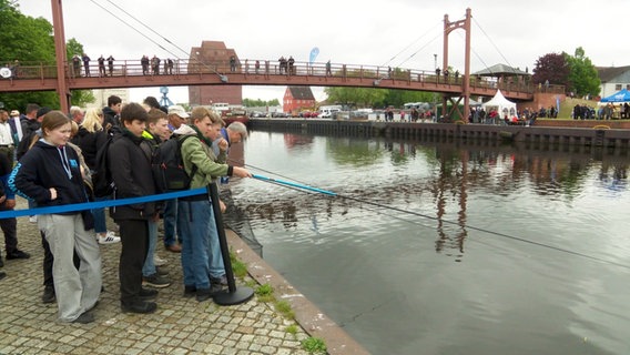 Viele Jugendliche stehen am Wasser, ein Junge hält eine Angel in der Hand. © Screenshot 