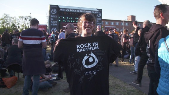 Eine Frau steht vor einer Bühne hält ein unterschriebenes Rock in Rautheim T-Shirt hoch. © Screenshot 