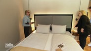 Zwei Frauen und ein Mann stehen neben einem Hotelbett. © Screenshot 