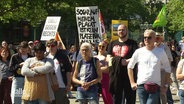 Mehrere Menschen haben sich in bunten Farben und mit Plakaten bei einer Kundgebung gegen die AfD auf einem Platz versammelt. © Screenshot 