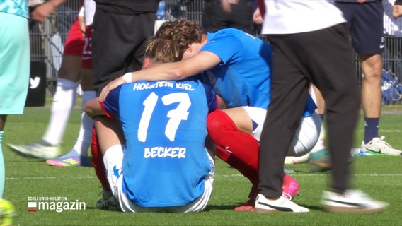Nach einem verlorenen Spiel sitzt ein Spieler des Vereins Holstein Kiel mit der Nummer 17 und dem Namen "Becker" auf dem Rasen und wird von einem Mitspieler getröstet. © Screenshot 