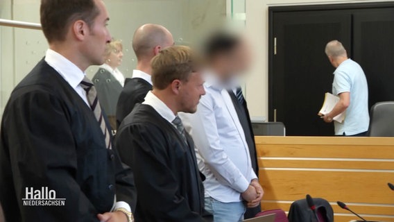 Ein Angeklagter steht neben seinen Anwälten. © Screenshot 