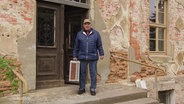 Ein älterer Mann steht vor einem alten Gutshaus. © Screenshot 