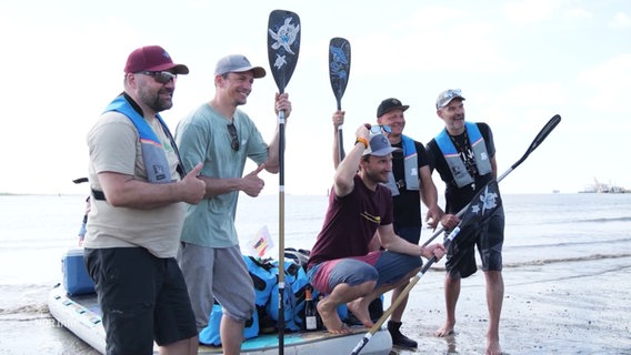 Fünf Männer stehen am Ufer, hinter ihnen ein großes Stand-up-Paddle Board. © Screenshot 