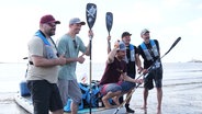 Fünf Männer stehen am Ufer, hinter ihnen ein großes Stand-up-Paddle Board. © Screenshot 