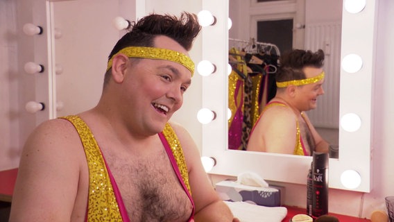 Ein Mann in einem knappen Glitzeroutfit sitzt in einer Künstlergarderobe und lächelt. Ein Mann in einem knappen Glitzeroutfit sitzt in einer Künstlergarderobe und lächelt. © Screenshot
