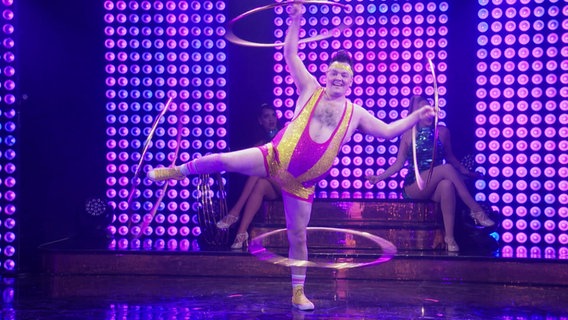 Ein Mann im knappen Glitzeroutfit steht auf einer Bühne und lässt an jedem seiner Arme und Beine einen Hula Hoop kreisen. © Screenshot 