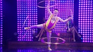 Ein Mann im knappen Glitzeroutfit steht auf einer Bühne und lässt an jedem seiner Arme und Beine einen Hula Hoop kreisen. © Screenshot 