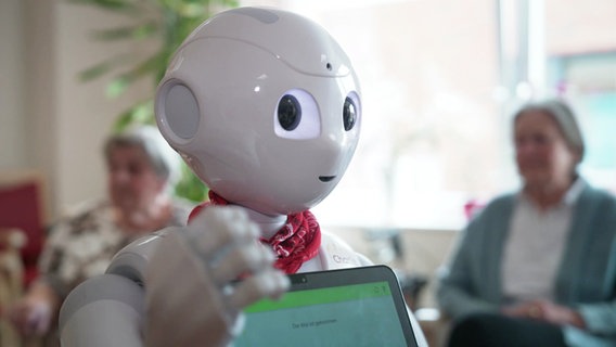 Ein niedlich aussehender Roboter mit Tablet vor der Brust steht vor zwei Seniorinnen die im Hintergrund sitzen. © Screenshot 
