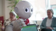 Ein niedlich aussehender Roboter mit Tablet vor der Brust steht vor zwei Seniorinnen die im Hintergrund sitzen. © Screenshot 