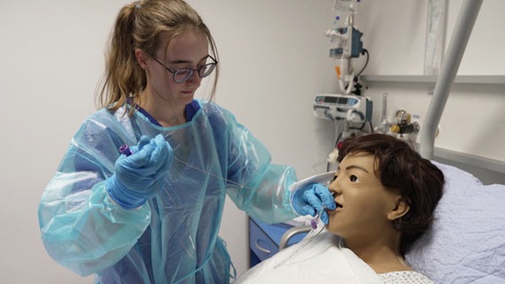 Eine junge Frau in Schutzkleidung führt eine medizinische Übung an einer realistisch aussehenden Pflegepuppe durch. Sie trägt blaue Handschuhe, Schutzbrille und Kittel und führt vorsichtig einen Schlauch in die Nase der Puppe ein. Die Szene findet in einem klinischen Übungsraum statt. © Screenshot 