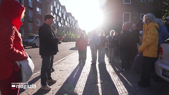Menschen bei einem Rundgang durch ein Stadtgebiet stehen im Sonnenuntergang vor einem Stolperstein. © Screenshot 