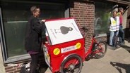 Ein rotes Lastenrad, links und rechts daneben stehen Personen. © Screenshot 