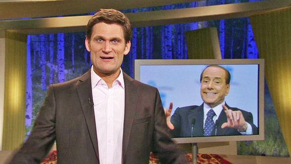 Moderator Christian Ehring neben einem Bild von Silvio Berlusconi.  