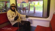 Michel Abdollahi in einem Altersanzug in der S-Bahn.  