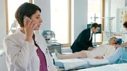 Eine junge Ärztin telefoniert, während sich im Hintergrund ein Mann und eine Krankenschwester zu einer Frau auf einem Krankenbett beugen. © NDR 