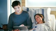 Ein junger Mann sitz auf der Bettkante einer jungen Frau im Krankenbett. Beide schauen auf ein Tablet.  