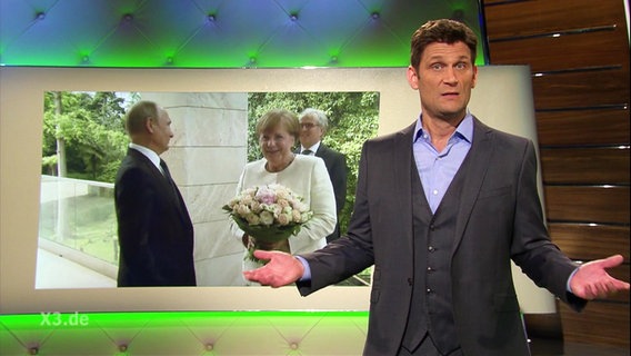 Christian Ehring moderiert extra 3, im Hintergrund ein Bild von Merkel und Putin.  