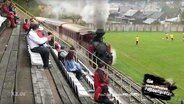 Eine Dampflock fährt direkt am Spielfeldrand eines Fußballplatzes  