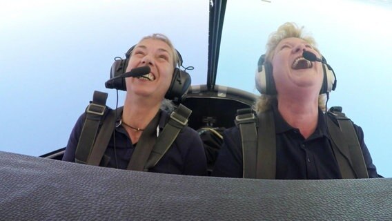 Zwei Frauen sitzen lachend im Cockpit eines Flugzeugs während eines Loopings.  
