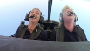 Zwei Frauen sitzen lachend im Cockpit eines Flugzeugs während eines Loopings.  