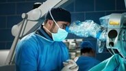 Dr. Matteo Moreau alias Mike Adler schaut durch ein OP-Mikroskop.  