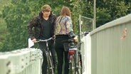 Zwei Fahrradfahrer versuchen an einer viel zu engen Stelle auf einer Brücke aneinander vorbeizukommen  