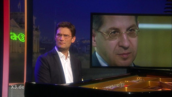 Christian Ehring sitzt am Klavier. Auf einem Bildschirm im Hintergrund ist der Kopf von Hans Georg Maaßen zu sehen.  