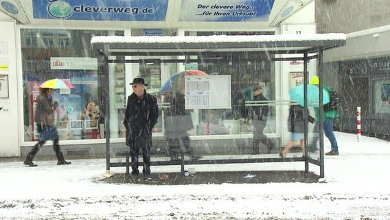 Ein Mann steht im dichten Schneetreiben unter dem Dach eines Wartehäuschens.  