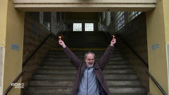 Ein Mann steht jubelnd und mit Lichterkerzen in beiden Händen vor einer langen Treppe, die als einziger Zugang zu einem Bahnsteig fungiert.  