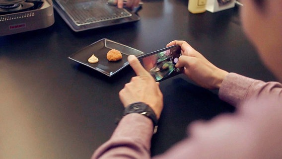 Auf einem Teller ist ein Hähnchen-Nugget aus Kunstfleisch angerichtet. Reporter Han Park fotografiert es mit seinem Smartphone.  
