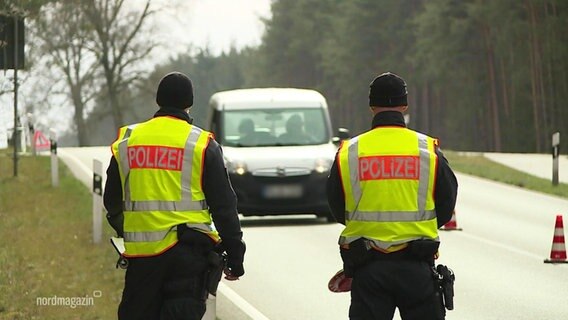 Zwei Polizisten stehen mit dem Rücken zur Kamera auf einer straße. Sie tragen gelbe Polizeiwarnwesten und ihnen kommt ein Pkw entgegen.  