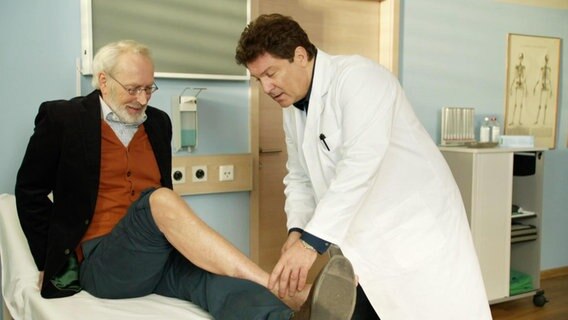 Dr. Christian Kleist (Francis Fulton-Smith) untersucht den Fuß eines Patienten, der auf einer Liege sitzt. © Screenshot 