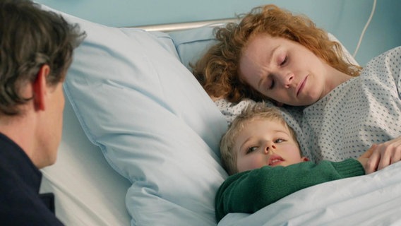 Die junge Mutter Miriam Reichardt liegt mit geschlossenen Augen in einem Krankenhausbett. Ihr Sohn Finn liegt neben seiner Mutter und spricht mit einem Mann, der am Bett sitzt.  