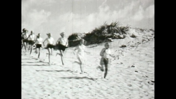 Schwarz-Weiß-Aufnahme von jungen Frauen, die hintereinander über den Strand Richtung Nordsee laufen.  