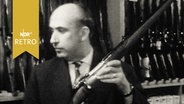 Waffenhändler Schlüter an seiner Ladentheke mit Jagdgewehr in der Hand  