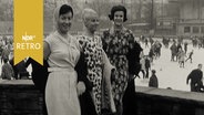 Drei Frauen posieren mit Kleidern vor einer Eisfläche mit Eisläufern  