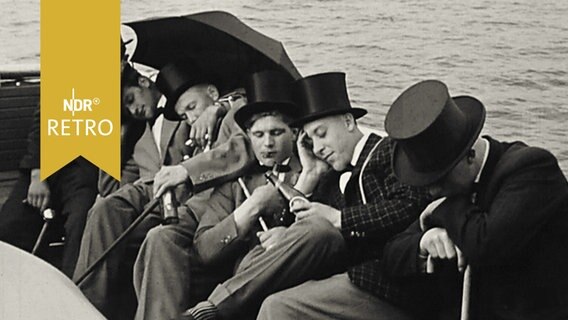 Sechs junge Männer mit Zylindern auf dem Kopf hängen halb schlafend an einer Bordwand auf See  