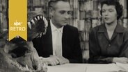 Geöffneter Raubfischkiefer auf einem Tisch neben zwei Personen beim Interview (1958)  