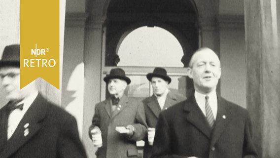 Vier mutmaßlich prominente Herren in Mänteln verlassen ein Gebäude mit Spendenbüchsen in der Hand (1959) zur Sammelaktion "Hamburg macht das Tor auf"  