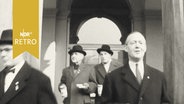 Vier mutmaßlich prominente Herren in Mänteln verlassen ein Gebäude mit Spendenbüchsen in der Hand (1959) zur Sammelaktion "Hamburg macht das Tor auf"  