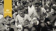 Zwei Studenten singen und musizieren mit Banjos inmitten einer Menge sitzender Jugendlicher.  