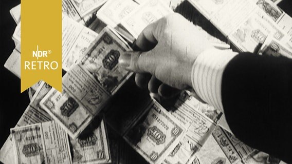 Eine Hand greift sich aus einem Haufen gebündelter DM-Scheine ein Bündel mit 10-Markscheinen heraus (1963)  