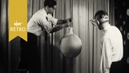 Zwei Männer Hängen in einer Möbelausstellung eine Lampe auf (1961).  