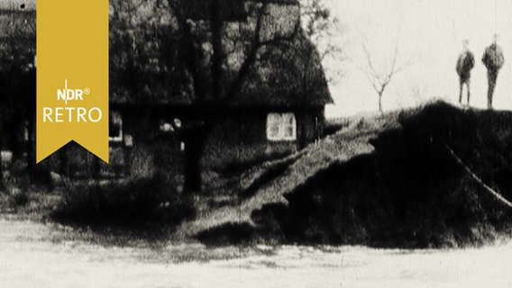 Gebrochener Deich: Zwei Männer stehen auf der Deichkrone, ein Häuser im Hintergrund werden vom Wasser umspült (1965)  