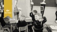 Erstklässer auf ihren Plätzen in einem Klassenraum halten ihre Schultüten hoch (1962)  